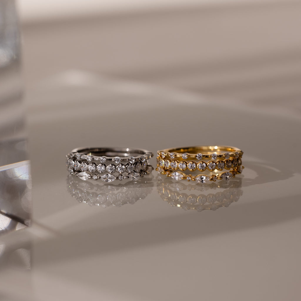 The Crystal Stacking Ring Set shown with one silver set of 3 rings and one gold set of 3 rings —adorned with crystal gemstones and displayed on a glossy, reflective surface.