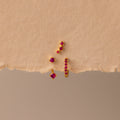 The Ruby Earring Trio Set, featuring delicate red gemstones, rests on a textured beige surface and displays its elegant charm.