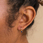Close-up of a person’s ear wearing Blue Zircon Drop Huggies—small gold earrings with a blue gemstone pendant, ideal for birthstone earring and personalized jewelry lovers.