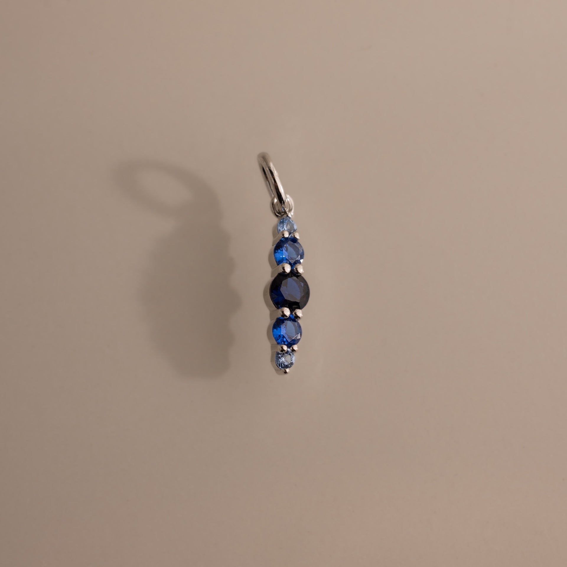 The Ombre Lace Sapphire Charm is a silver pendant featuring four vertically arranged blue gemstones, making a stunning birth month gift or personalized charm, shown on a plain beige background.