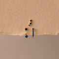 The Sapphire Earring Trio Set features four gold birthstone earrings with small blue gemstones, including a pavé huggie hoop, displayed on a textured beige surface.