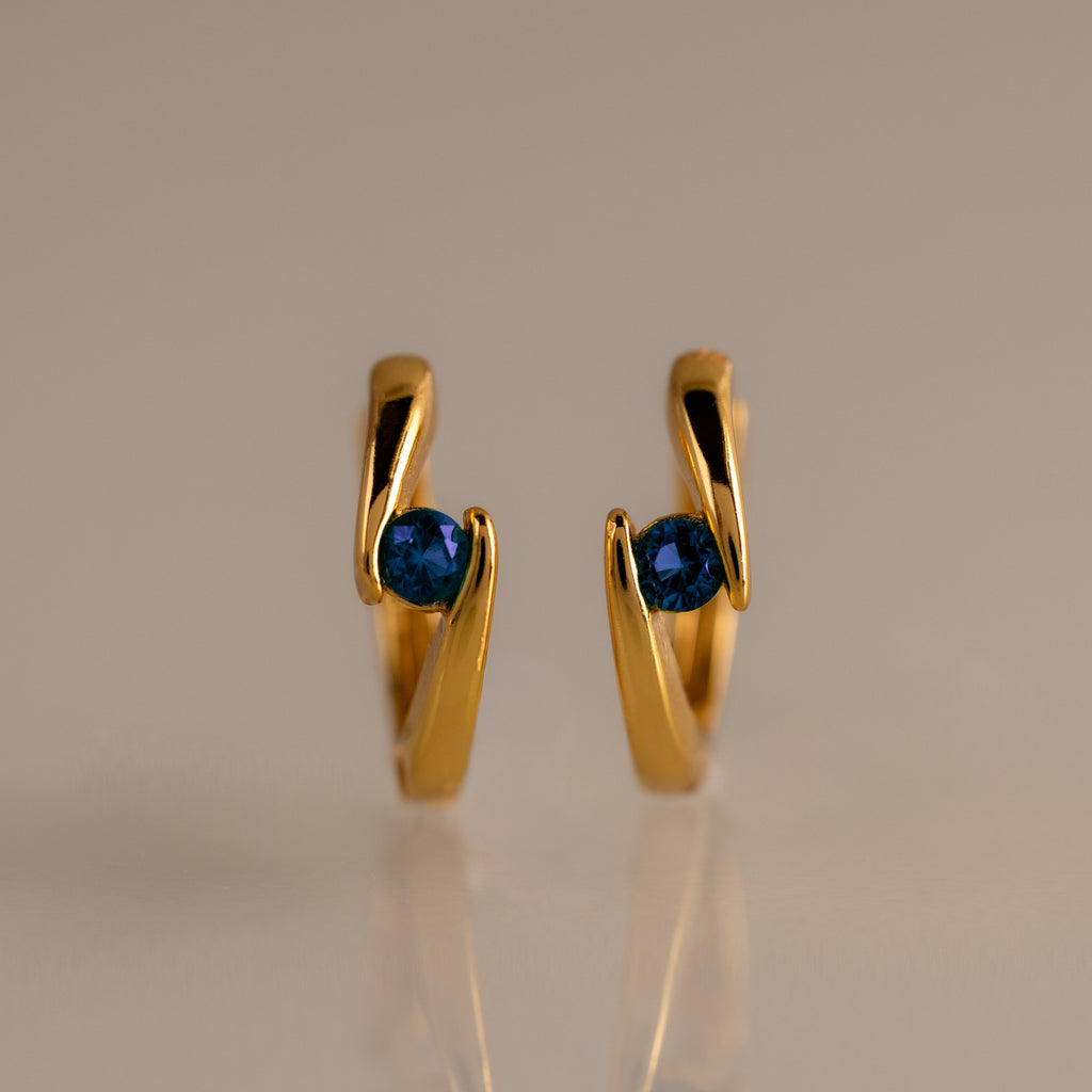 Floating Sapphire Huggies—gold hoop earrings with blue gemstones—stand upright on a neutral, reflective surface, making them the perfect personalized jewelry piece.