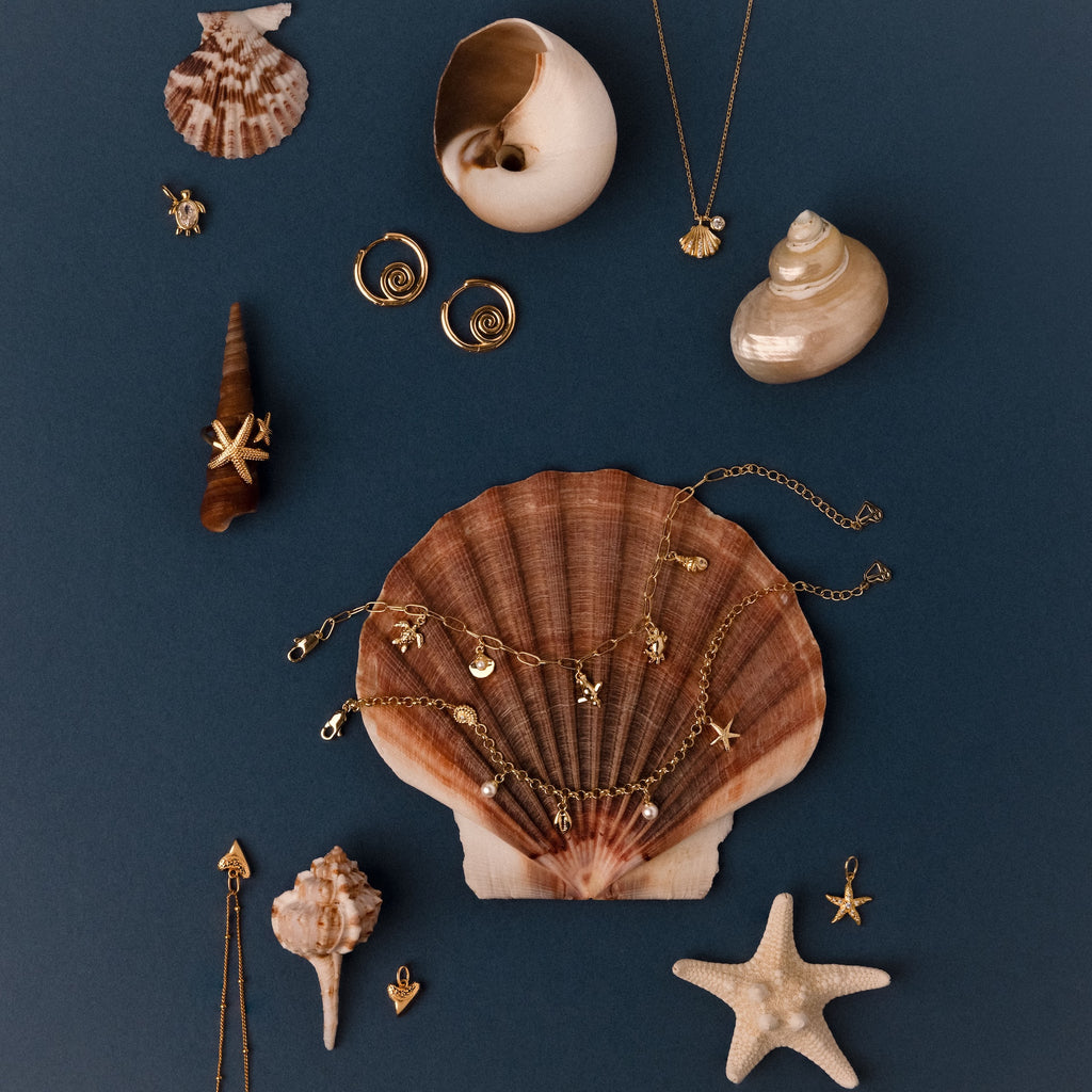 The Seaside Pearl Charm Bracelet and other ocean-inspired gold coastal jewelry, like necklaces and seashells, are displayed on a dark blue background.