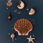 The Seaside Pearl Charm Bracelet and other ocean-inspired gold coastal jewelry, like necklaces and seashells, are displayed on a dark blue background.