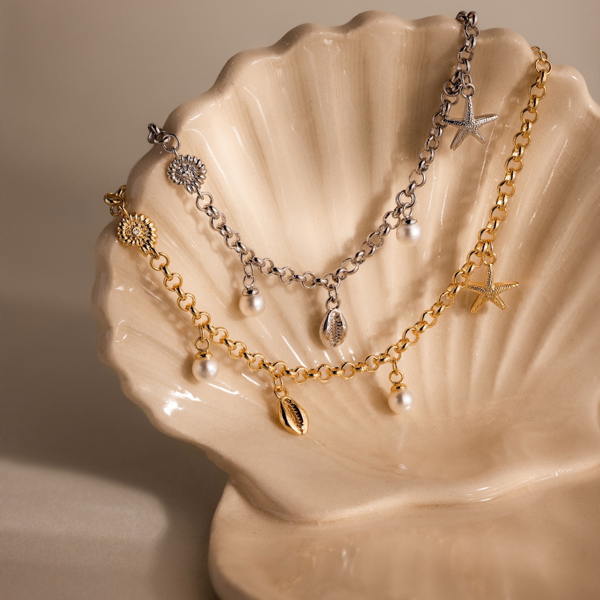 Gold and silver coastal jewelry with ocean-inspired charms is displayed on a beige shell dish. Add the Seaside Pearl Charm Bracelet to your collection for a touch of seaside elegance.
