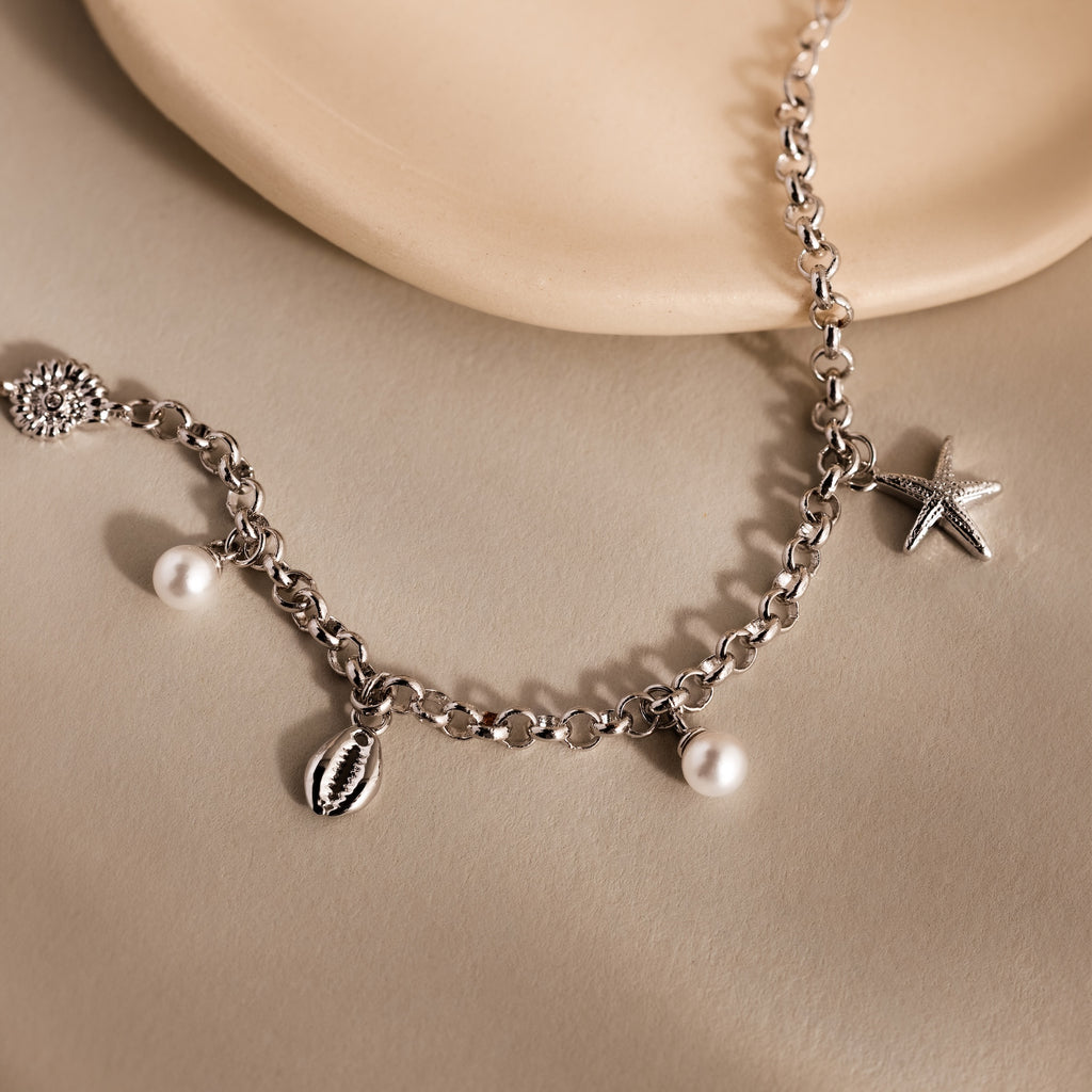 The Seaside Pearl Charm Bracelet in Silver features starfish, shell, and pearl charms on a delicate chain—perfect for coastal jewelry lovers. Displayed on a beige surface.