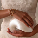 Woman in a textured white dress holding a seashell, adorned with coastal jewelry and the Seaside Pearl Charm Bracelet featuring ocean-inspired charms.