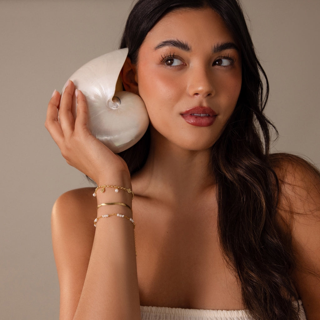 A woman with long dark hair in a white top holds a large seashell to her ear, wearing the Seaside Pearl Charm Bracelet—coastal jewelry adorned with ocean-inspired charms.