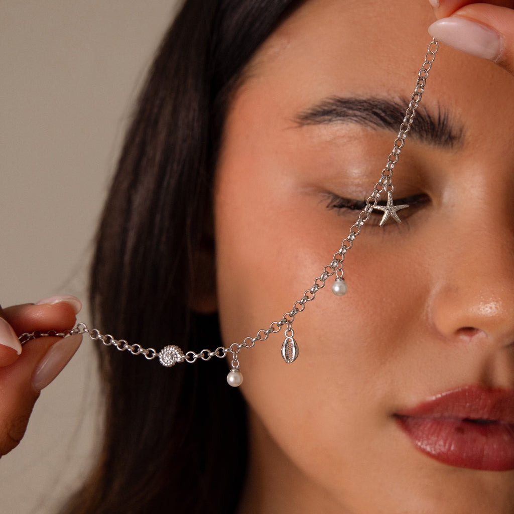 A woman holds the Seaside Pearl Charm Bracelet in Silver—an elegant coastal jewelry piece with pearls and a starfish, inspired by the ocean, in front of her closed eyes.
