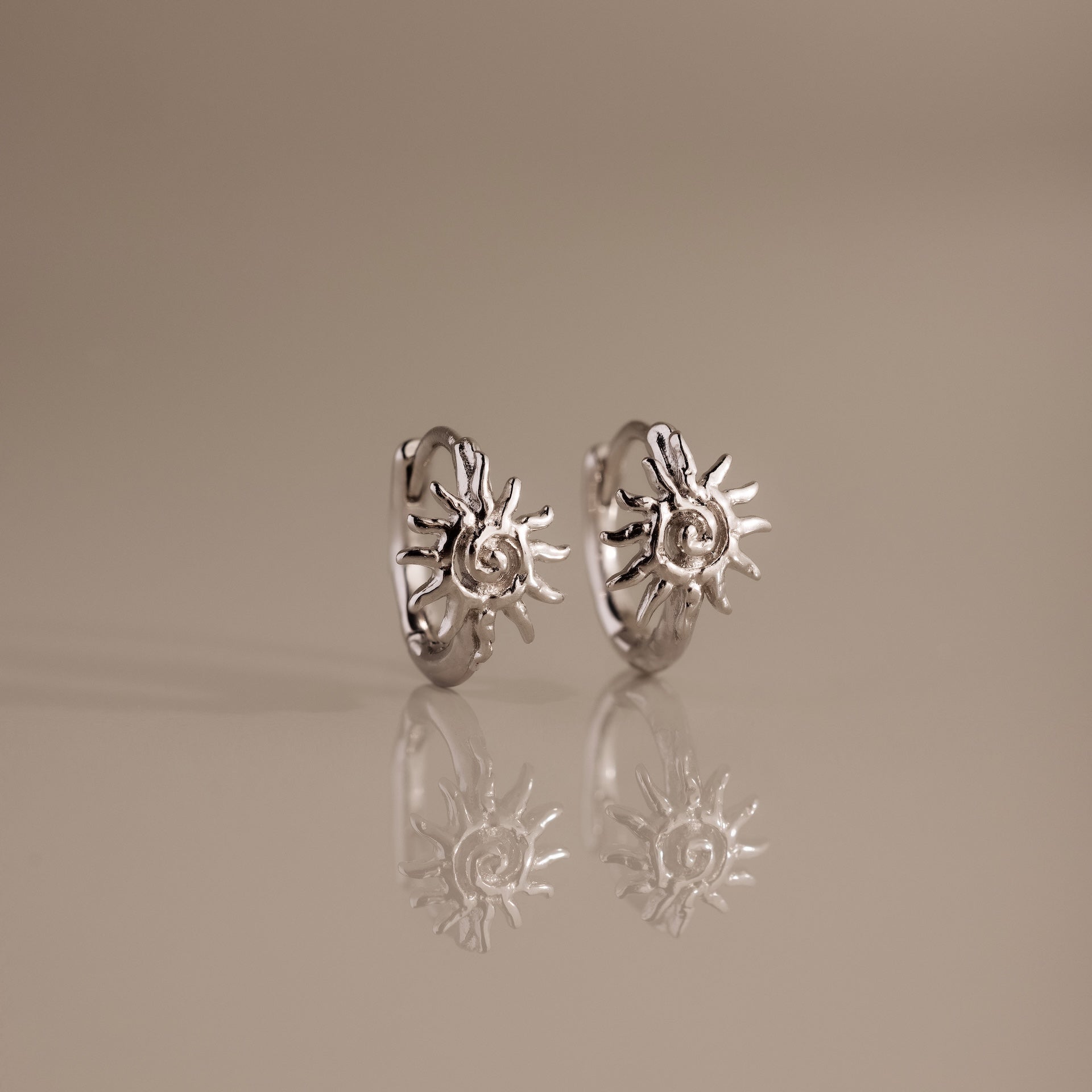 Celestial Sun Huggies, a pair of silver sunburst earrings, stand on a reflective beige surface—ideal statement jewelry for a radiant touch.