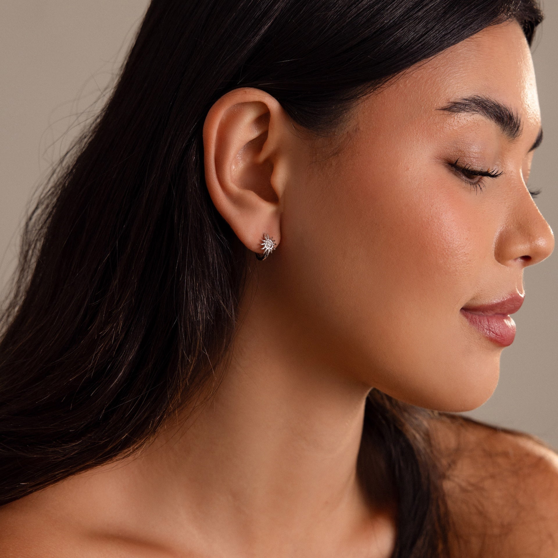 A woman with long dark hair, eyes closed in profile, wears the Celestial Sun Huggies in Silver—small star-shaped earrings that radiate subtle, statement charm.