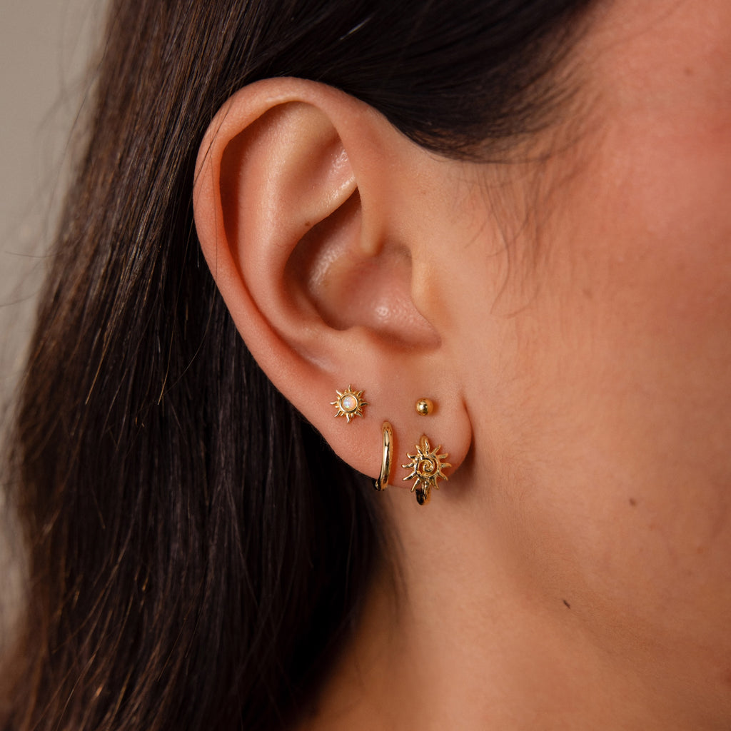Close-up of an ear with three gold earrings: a small sun stud, Celestial Sun Huggies featuring a sun charm, and a tiny gold stud—ideal for statement jewelry lovers.