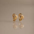 Celestial Sun Huggies in 18K Gold feature sun-shaped designs and are perfect statement earrings for any occasion, shown reflected on a smooth, neutral surface.