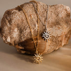 Two Celestial Sun Necklaces, each with a sunburst pendant on gold and silver chains, are elegantly displayed on a textured rock.