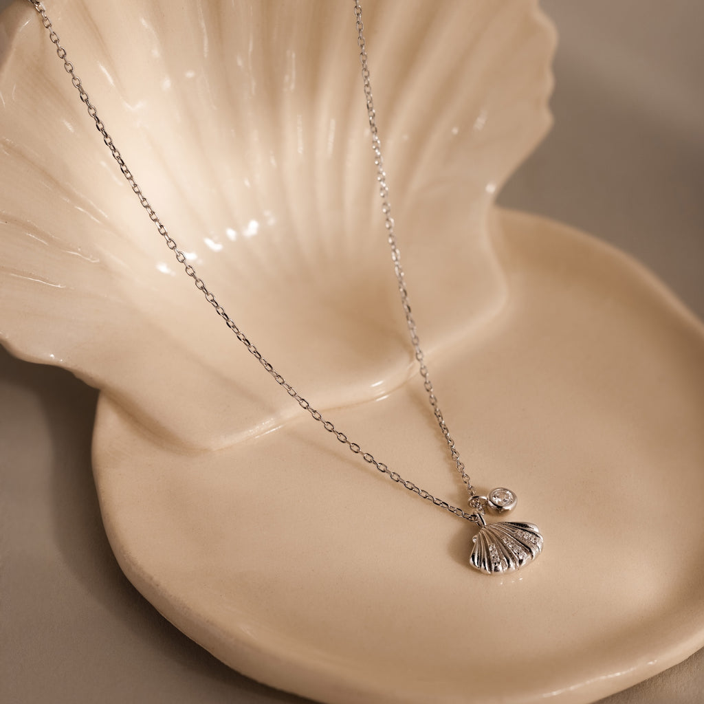 The Diamond Seashell Charm Necklace in Silver, featuring a delicate shell pendant and sparkling gem, is an elegant ocean-inspired piece displayed on a cream ceramic shell dish.