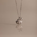 The Diamond Seashell Charm Necklace in Silver features a shell pendant and a small round gemstone, displayed on a neutral background—an elegant accessory to elevate any look.