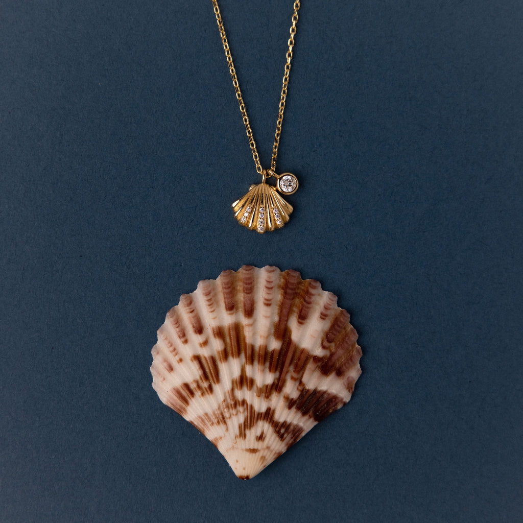 The Diamond Seashell Charm Necklace in 18K Gold showcases a shell pendant with a sparkling gem above a brown and white seashell, reflecting coastal elegance.