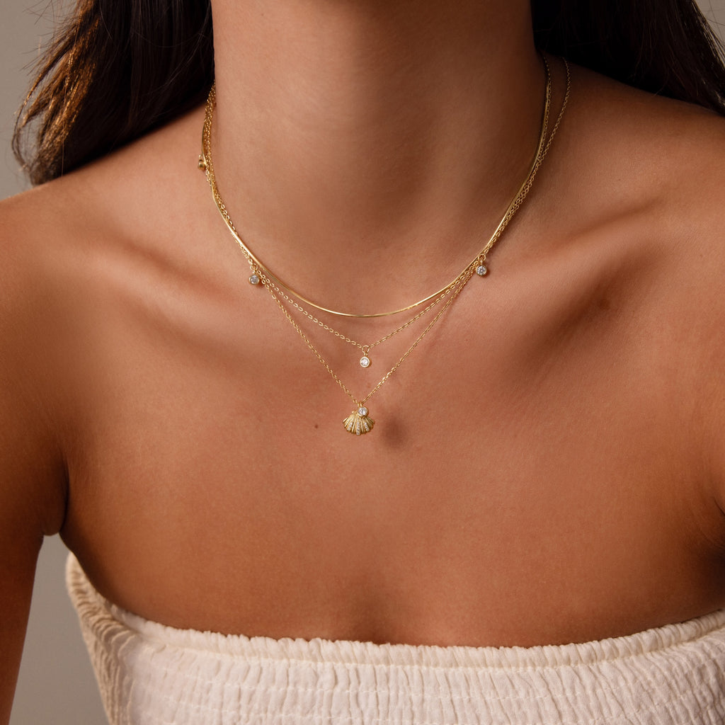 Woman shown from the shoulders up wearing a white, strapless textured top and layered gold necklaces, including the Diamond Seashell Charm Necklace in 18K Gold.