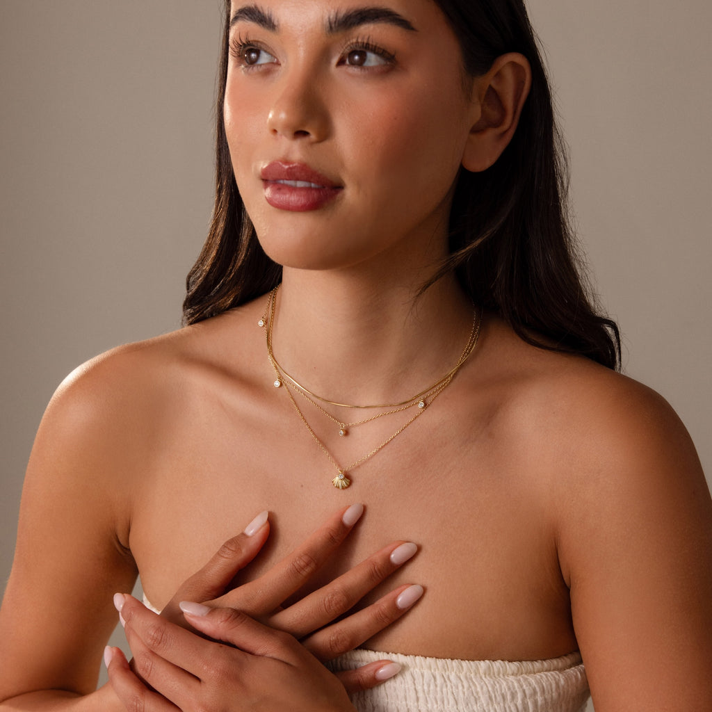 A woman in a strapless top gently touches her chest, showcasing layered gold necklaces, including the Diamond Seashell Charm Necklace in 18K Gold.