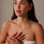 A woman with long dark hair wears a strapless top and layered gold necklaces, featuring the Diamond Seashell Charm Necklace, hands clasped near her chest.