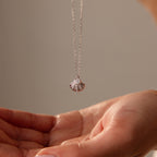 A hand reaches for the Diamond Seashell Charm Necklace in Silver, featuring a shell pendant with a sparkling gemstone on a delicate chain.