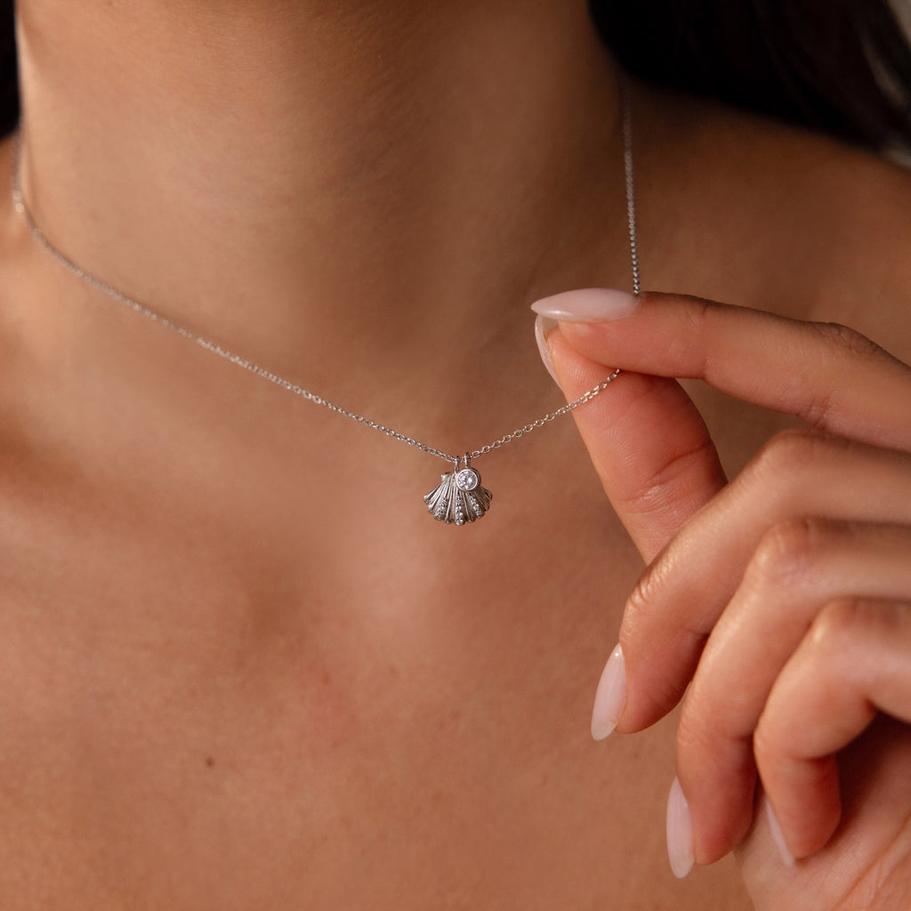 A woman holds the Diamond Seashell Charm Necklace in Silver with a shell pendant close to her neck, displaying manicured nails.