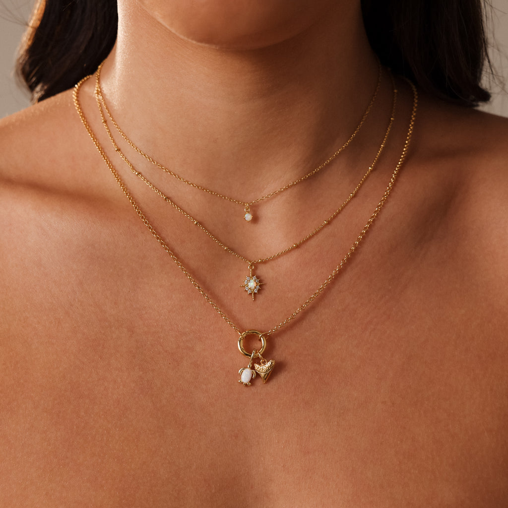 Woman wearing three layered gold necklaces with small pendants, including a flower, a pearl, and an ocean-inspired Opal Sea Turtle Charm in Silver for a seaside jewelry touch.