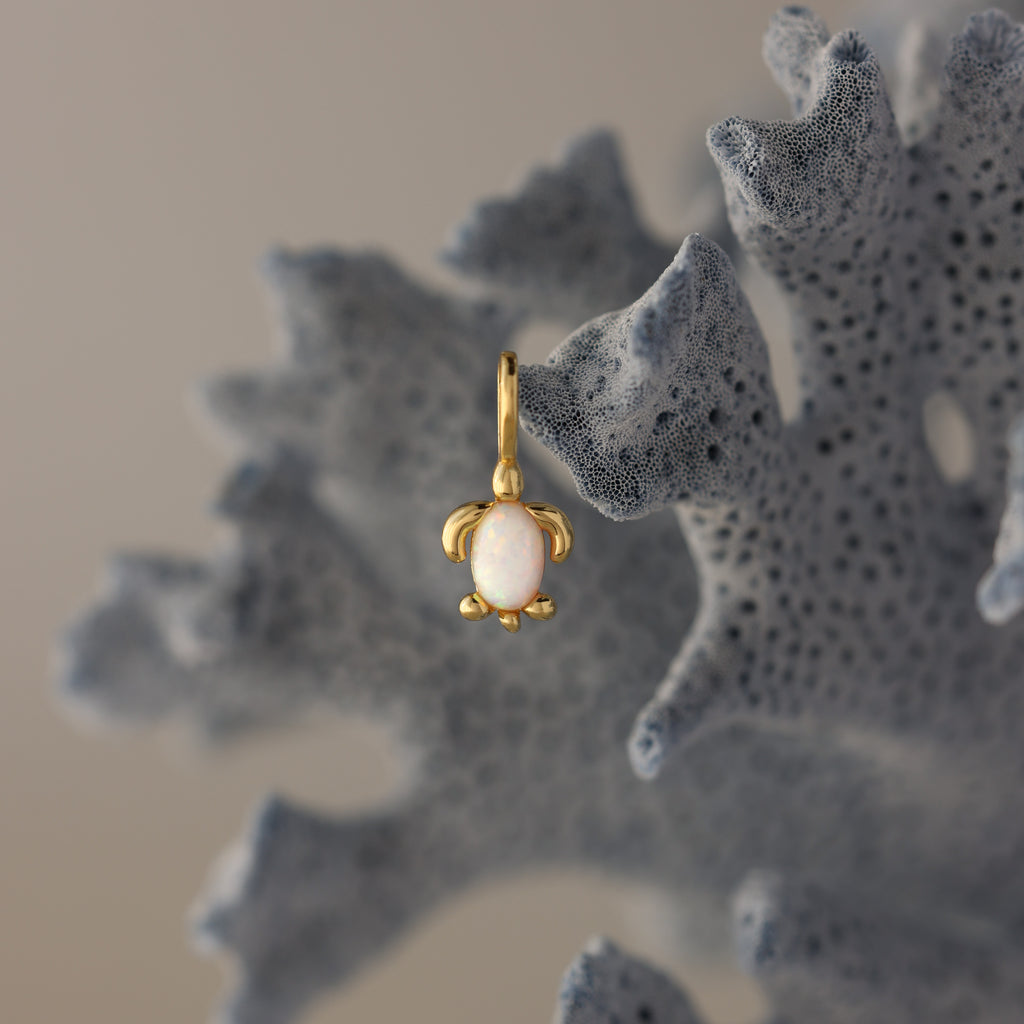 The Opal Sea Turtle Charm in 18K Gold, featuring a white stone and turtle design, hangs on textured blue coral for an ocean-inspired accent.