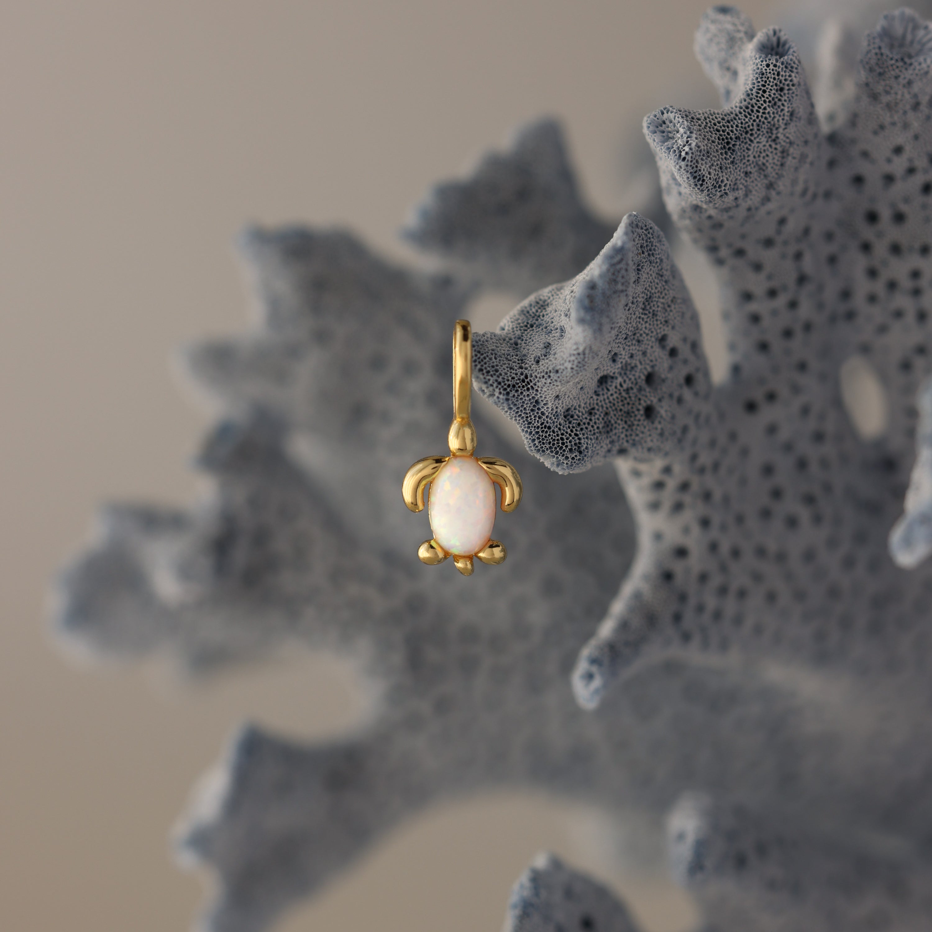 The Opal Sea Turtle Charm in 18K Gold, featuring a white stone and turtle design, hangs on textured blue coral for an ocean-inspired accent.
