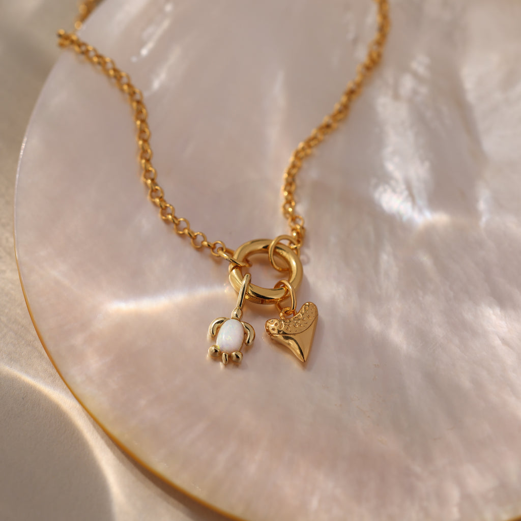 Opal Sea Turtle Charm in 18K Gold features a gold chain necklace with pearl, heart, and sea turtle charms on an iridescent shell surface for an ocean-inspired look.
