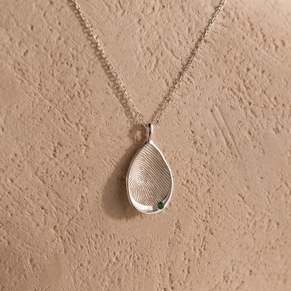 The Organic Fingerprint Teardrop Necklace in Sterling Silver features a textured teardrop and green gem on a delicate chain—a unique, personalized keepsake shown against a beige background.