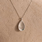 The Organic Fingerprint Teardrop Necklace in Sterling Silver features a textured teardrop and green gem on a delicate chain—a unique, personalized keepsake shown against a beige background.