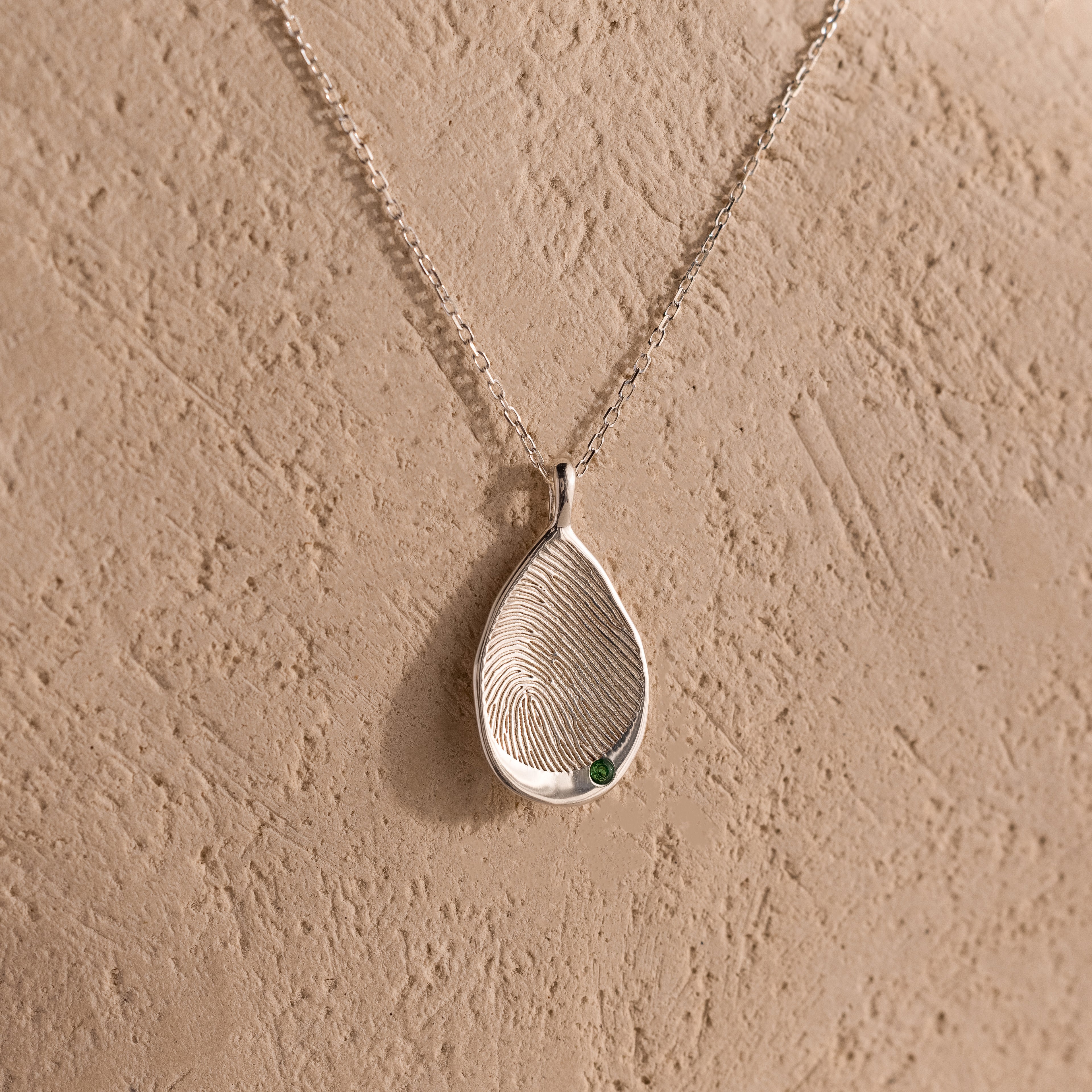 The Organic Fingerprint Teardrop Necklace in Sterling Silver features a textured teardrop and green gem on a delicate chain—a unique, personalized keepsake shown against a beige background.