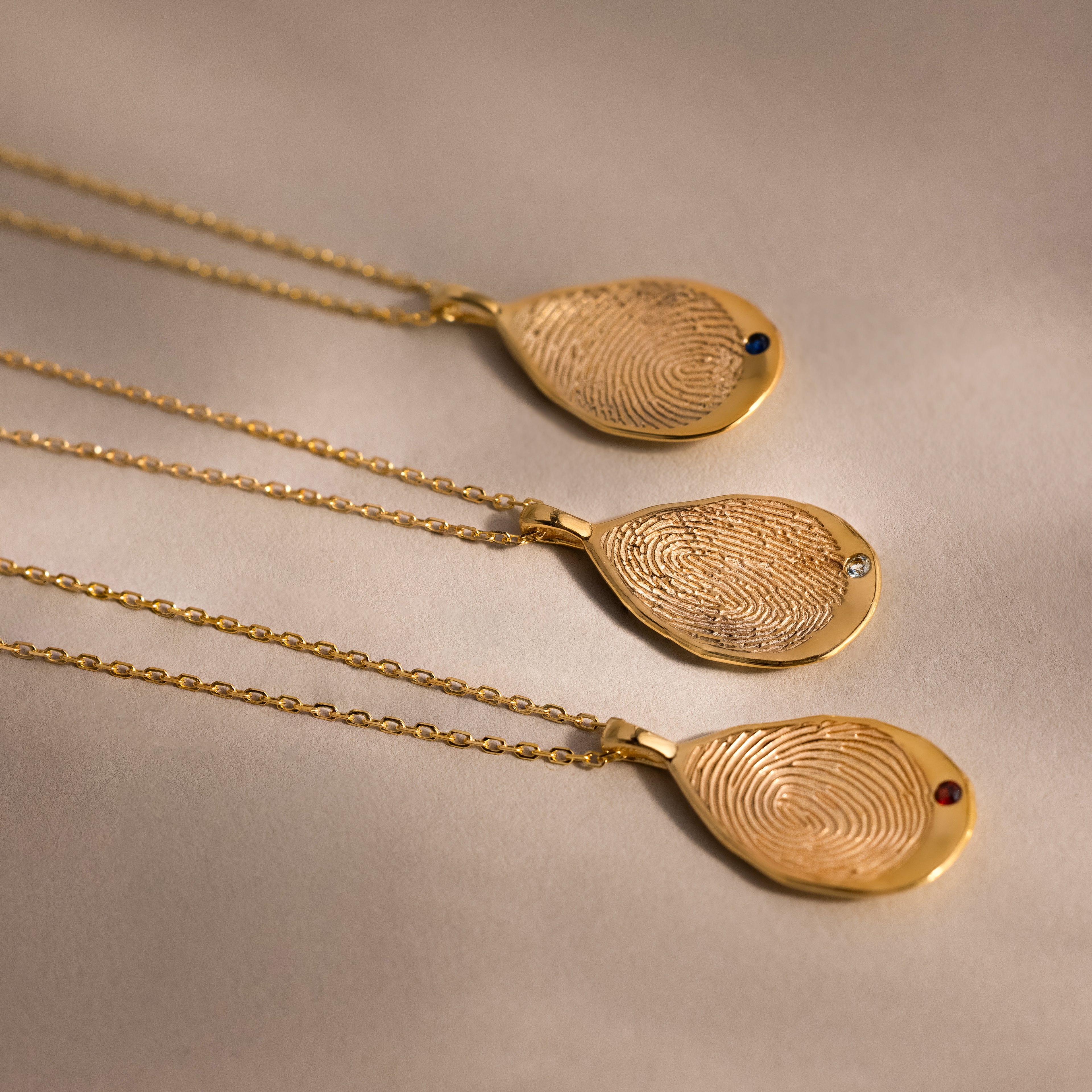 Three Organic Fingerprint Teardrop Necklaces in 18K Gold, each with unique fingertip patterns and small birthstone gems, create a personal keepsake, elegantly displayed on a beige surface.