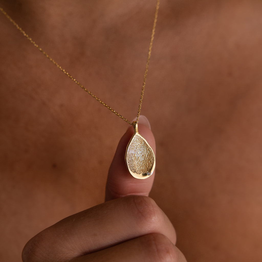 A person holding an Organic Fingerprint Teardrop Necklace, beautifully crafted as a gold teardrop pendant and engraved with a unique fingerprint.