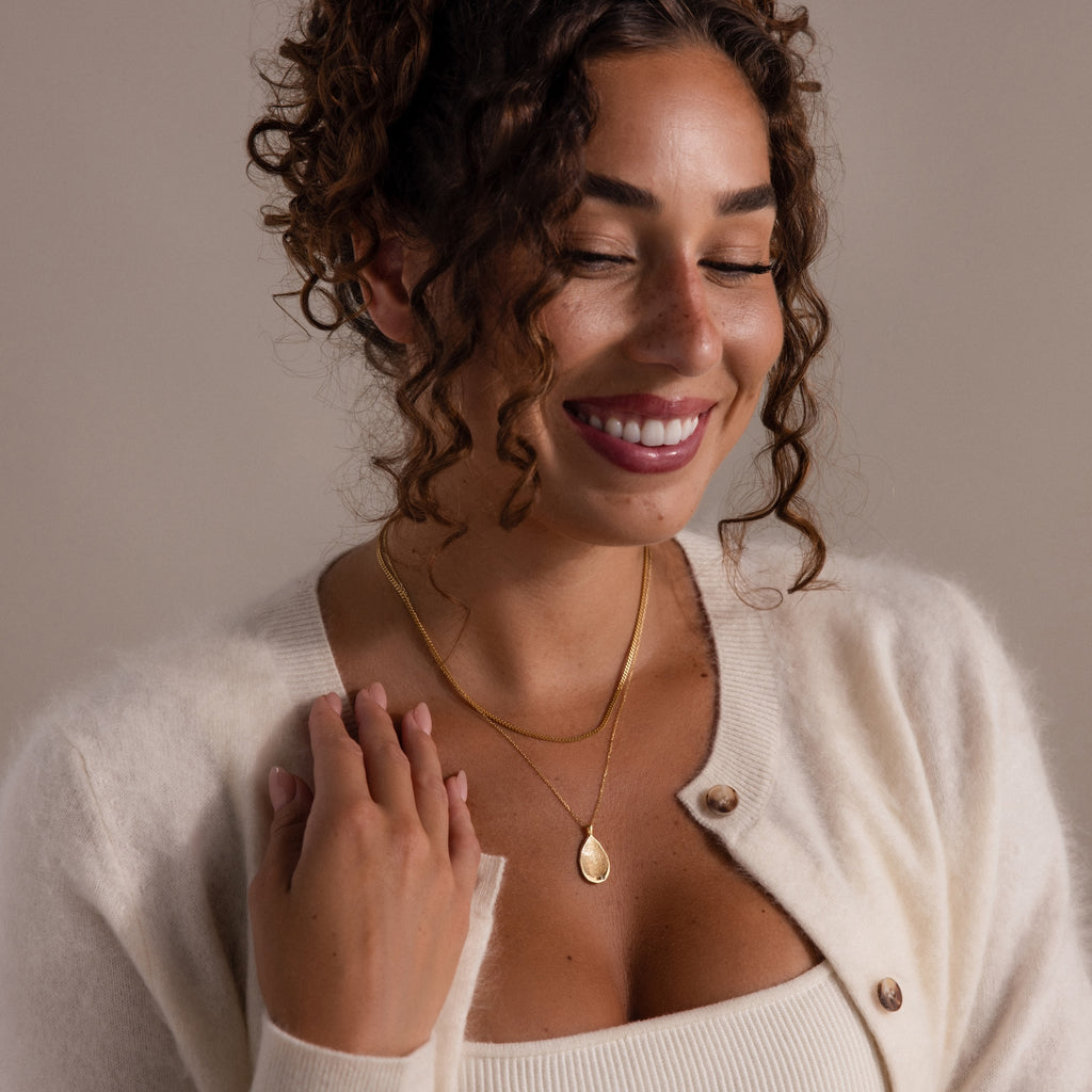 A woman with curly hair smiles in a cream sweater, showcasing the Organic Fingerprint Teardrop Necklace layered with gold chains against a neutral background.