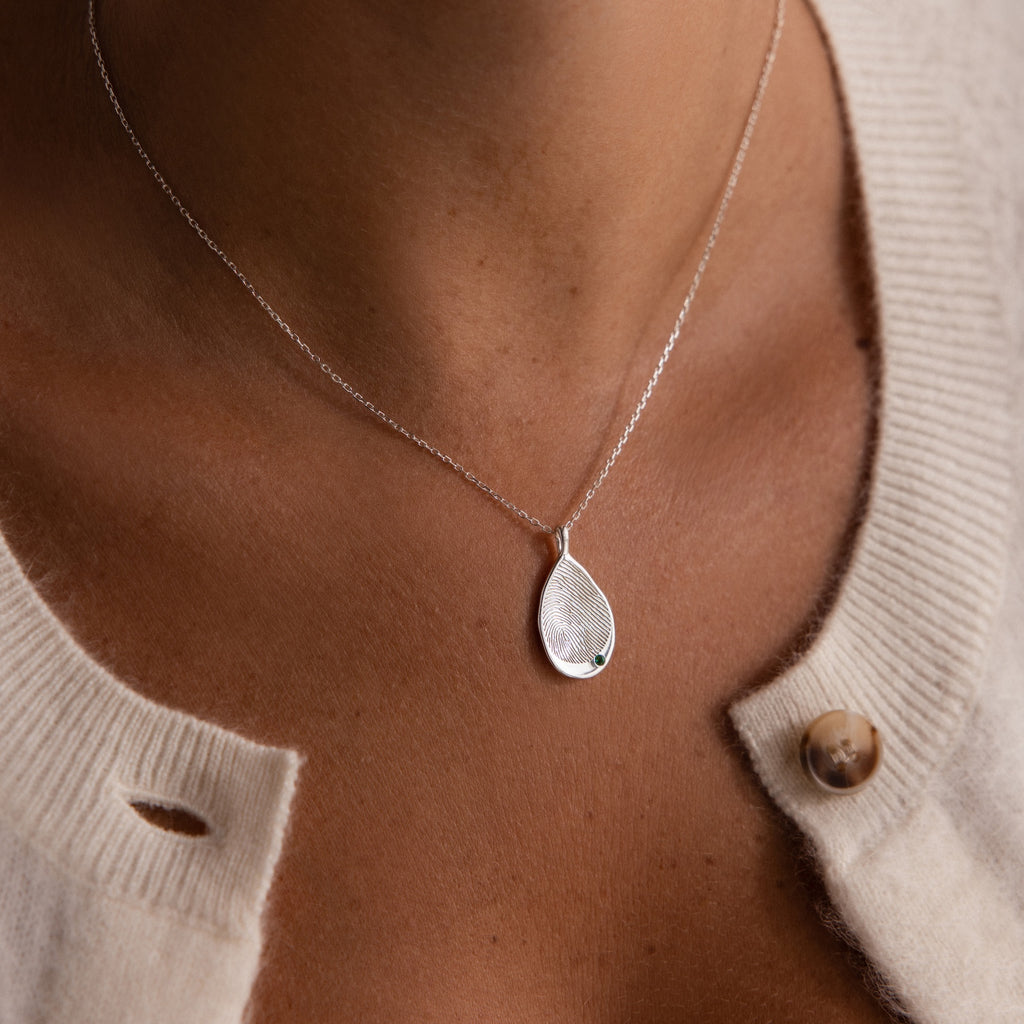 A personalized keepsake, the Organic Fingerprint Teardrop Necklace in Sterling Silver features a small green gem, making it an elegant birthstone accessory worn beautifully over a cream sweater.