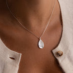 A personalized keepsake, the Organic Fingerprint Teardrop Necklace in Sterling Silver features a small green gem, making it an elegant birthstone accessory worn beautifully over a cream sweater.