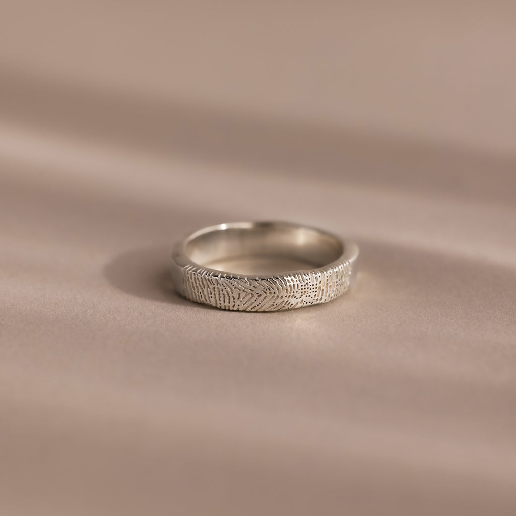 The Organic Fingerprint Band in Sterling Silver, featuring a delicate leaf pattern, rests on a soft beige surface—ideal as a memorial keepsake or custom-engraved ring for treasuring special memories.