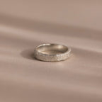 The Organic Fingerprint Band in Sterling Silver, featuring a delicate leaf pattern, rests on a soft beige surface—ideal as a memorial keepsake or custom-engraved ring for treasuring special memories.