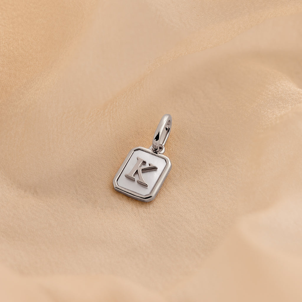 A silver square pendant charm with a raised letter 'K' on a soft beige fabric background.