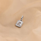 A silver square pendant charm with a raised letter 'K' on a soft beige fabric background.