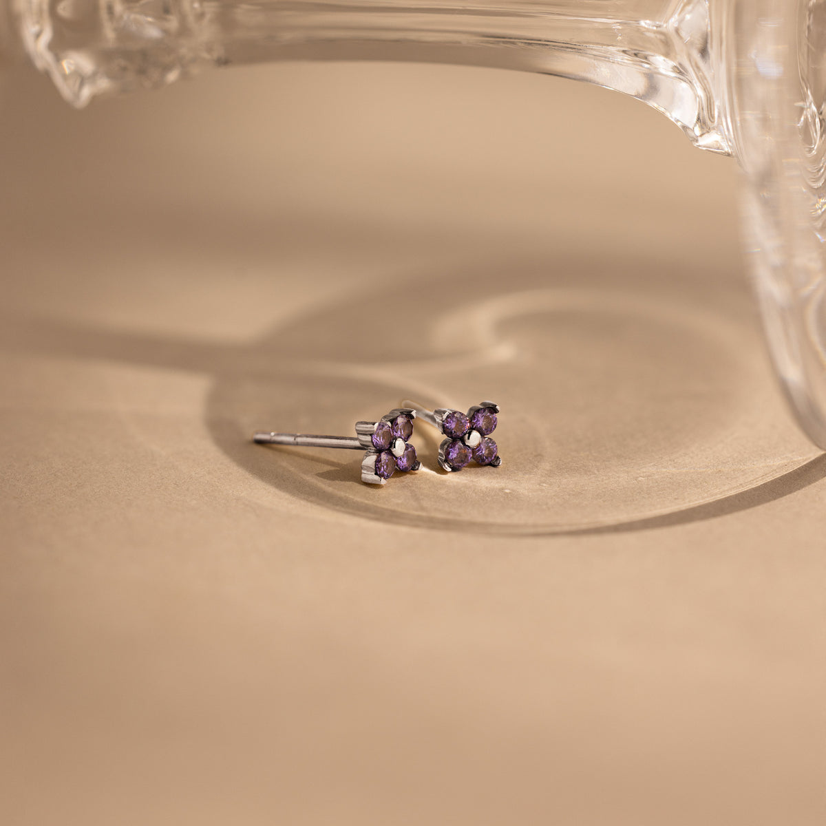 A pair of small silver stud earrings with four purple gemstones arranged in a flower shape, resting on a beige surface with a glass object in the background.
