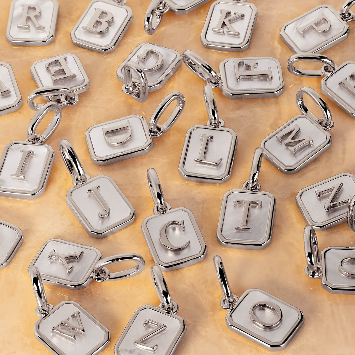 A close-up of multiple silver letter charms  arranged on a warm beige surface. Each charm is a rounded square with an embossed capital letter and a small jump ring clasp.