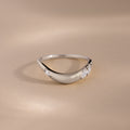 A silver teardrop-shaped ring with three clear gemstones, photographed on a warm beige background.