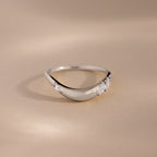 A silver teardrop-shaped ring with three clear gemstones, photographed on a warm beige background.