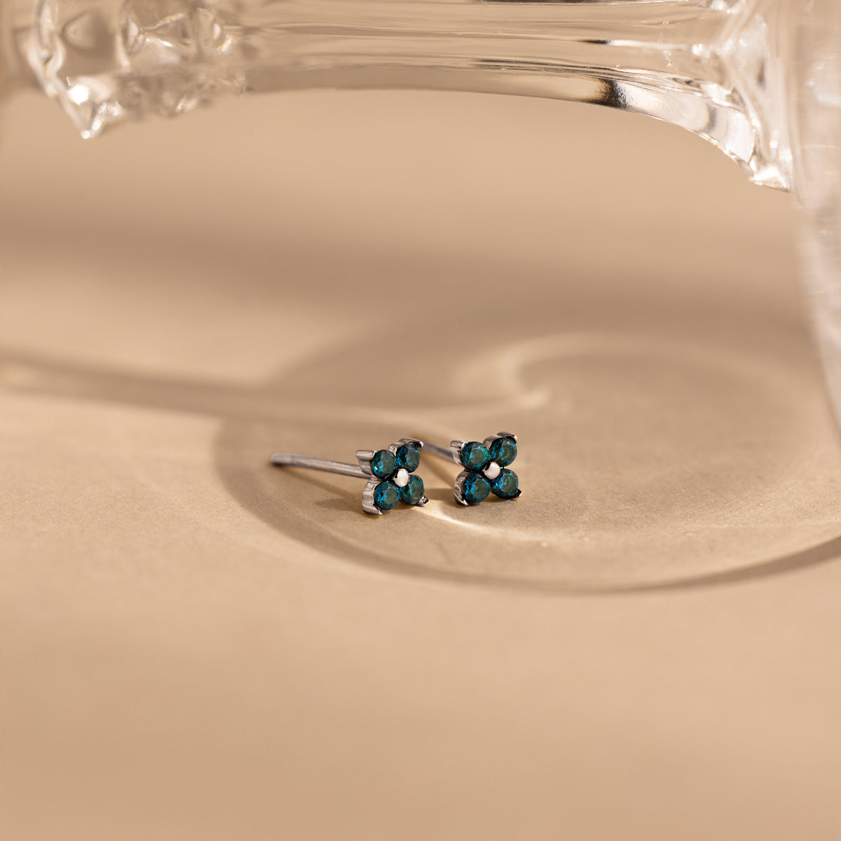 A pair of small silver stud earrings with four dark teal-blue gemstones arranged in a flower shape, resting on a warm beige surface with a glass object partially visible at the top.