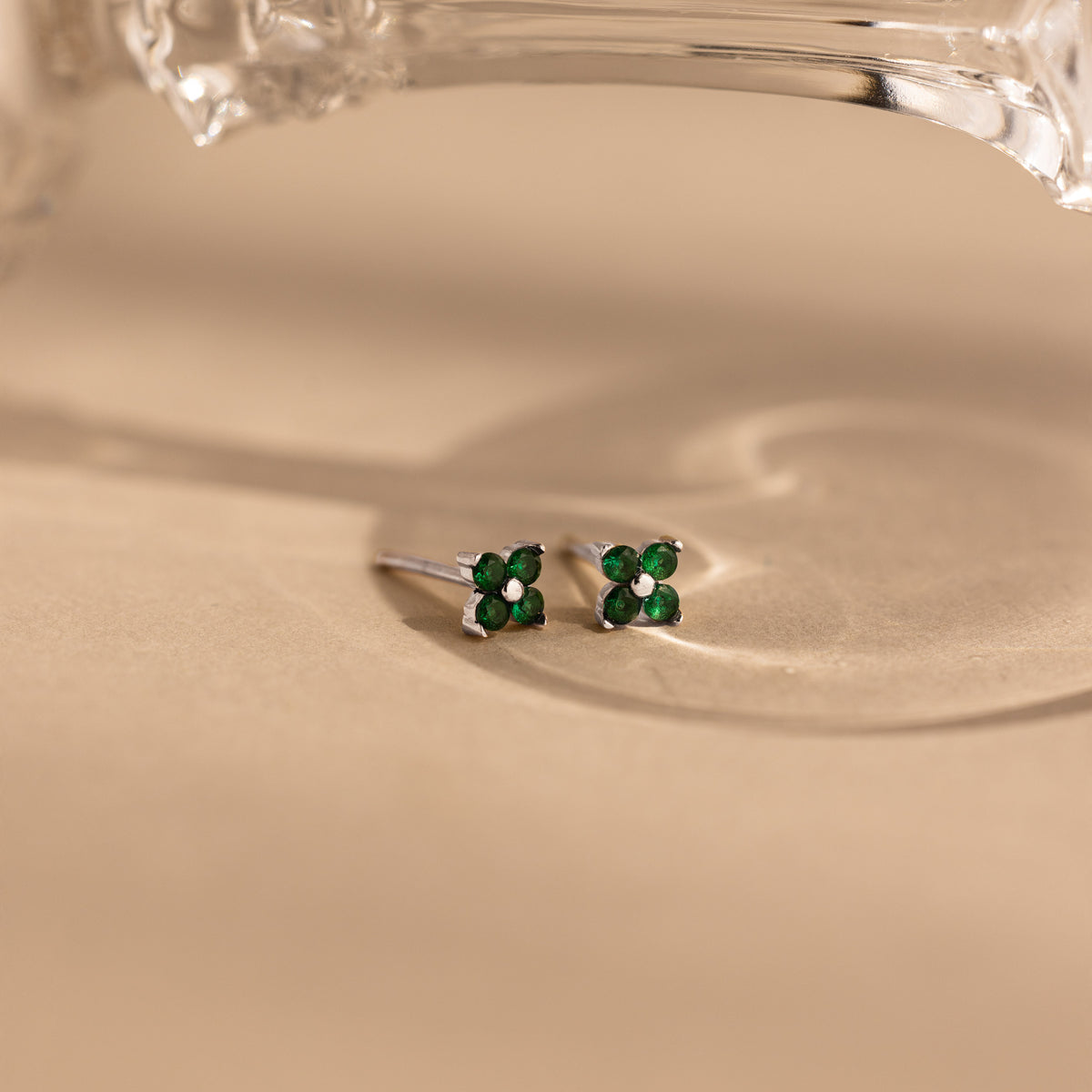 A pair of small silver stud earrings with four green gemstones arranged in a flower shape, resting on a warm beige surface with a soft shadow and part of a clear glass object at the top.