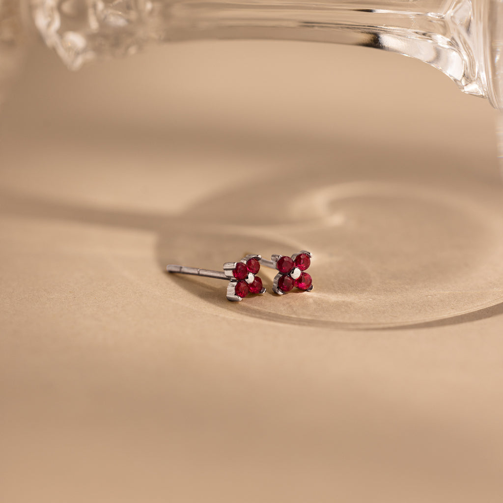 A pair of small silver stud earrings with four pink gemstones arranged in a flower shape, resting on a warm beige surface with a soft shadow and part of a clear glass object at the top.