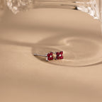 A pair of small silver stud earrings with four pink gemstones arranged in a flower shape, resting on a warm beige surface with a soft shadow and part of a clear glass object at the top.